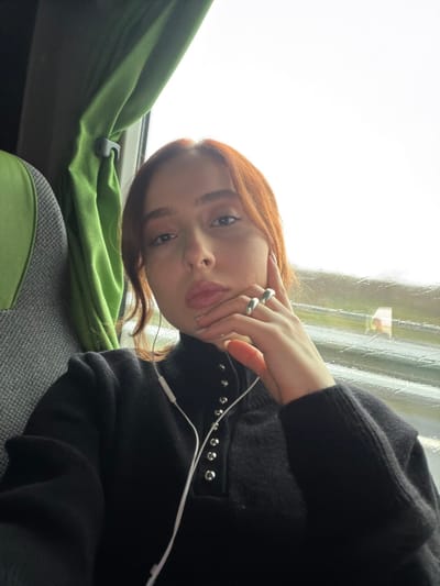 The image captures a young individual, female, with red-brown hair, seated within a vehicle, reportedly in Moineville, France. She is wearing a black long-sleeved top featuring a mock-neck and a front placket adorned with silver-colored eyelets or snaps. White wired earphones are visible around her neck and leading to her ears. Her left hand is positioned near her chin, displaying two silver-colored rings on her ring finger. To her left, part of a grey fabric seat and a green curtain are visible. To her right, a window shows a blurred exterior with an overcast sky and streaks of rain or moisture on the glass.