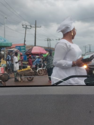 A medium shot captures a street scene in Aketu-Oja, Nigeria, viewed from inside a vehicle. In the foreground, a dark dashboard and a cracked windshield are visible. The middle ground presents a bustling market with several people and vehicles. A woman in a white dress and headscarf, wearing large gold earrings, is prominently featured on the right, holding a book or pamphlet. To her left, a man in a white shirt stands near a cart loaded with yellow fruits. Several other individuals, some on motorcycles, are dispersed throughout the market, with stalls and awnings suggesting a commercial area. The background reveals power lines and overcast skies, indicating an outdoor, possibly urban, setting.