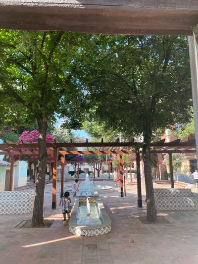 This outdoor scene, set in Picanya, Spain, captures a sunny day in a public plaza or park. The image displays a long, inviting pathway paved with light brown and terracotta-colored tiles, extending into the distance under a bright blue sky.

Prominently featured is a decorative, rectangular fountain with multiple water jets, its sides adorned with ornate blue, white, and yellow/gold patterned tiles. Two children are visible near the fountain; one, wearing a striped shirt and shorts, stands beside a yellow ball in the foreground, while another child in a light top is further down the fountain's length.

Along the pathway, a sturdy wooden pergola structure provides shade, partially covered by lush green foliage and vibrant pink flowering plants, likely bougainvillea. Large, leafy trees with substantial trunks are spaced along the path, contributing to the shaded ambiance. Beyond the pergola and trees, various buildings with light-colored walls and tiled elements are visible, characteristic of Mediterranean architecture.

The bright sunlight, evident from the distinct shadows cast on the pavement, suggests it is midday or early afternoon. The overall atmosphere appears peaceful and leisurely, with people enjoying the pleasant outdoor setting. No discernible text is visible.