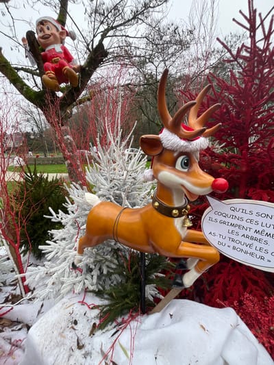 The image captures an outdoor festive display in Bagnoles de l'Orne Normandie, France, under an overcast sky. A large, brown, cartoon-style reindeer figure, featuring a red nose, antlers, a Santa hat, and a brown collar with gold bells, is depicted in a leaping pose, supported by a dark pole. A white speech bubble originating from the reindeer's mouth displays French text. To the left, a red-suited elf figure with a red and white hat is perched in the bare, moss-covered branches of a large deciduous tree. The foreground and midground are adorned with several artificial white-flocked Christmas trees, vibrant red artificial trees or branches, and patches of artificial snow covering the ground, interspersed with natural green foliage and scattered red twigs. The background consists of a park-like environment with bare trees and distant green areas.