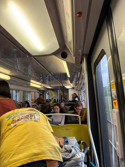 The interior of a train car in La Courneuve, France (48.922, 2.378). The train car is filled with passengers seated on yellow-padded benches. A man in a yellow shirt with a cartoon image of a food cart printed on it is standing facing the rear of the train car, facing the camera. Another man, wearing a red shirt and a face mask is standing near the front of the train car. Several other passengers are seated facing forward, their faces mostly obscured. A window to the right of the frame shows a view of trees and shrubs. The train car has an orange button located on the ceiling. The floor is clean and tiled. The train car is well-lit with several overhead lights. A baby stroller sits in the lower right corner of the frame. The train is moving with a slight vibration. A sign near the front of the train car indicates a stop. The scene is calm and quiet.