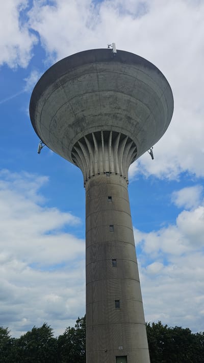 A low-angle, eye-level, medium outdoor shot captures a towering concrete water tower against a backdrop of a bright blue sky with scattered white clouds. The water tower has a large, rounded bowl-shaped top that tapers down to a cylindrical shaft. The shaft is segmented into horizontal bands, each with a textured, ribbed surface. Several small, rectangular windows are visible on the shaft. At the very top of the tower, on the rim of the bowl, a small antenna or communication equipment is mounted. Along the curved edge of the bowl, near the top of the shaft, two more pieces of equipment are attached, possibly also for communication or monitoring. The lower portion of the image shows a dark line of trees, indicative of a natural or park setting. The water tower is the dominant subject, situated in the city of Silenrieux, Belgium.