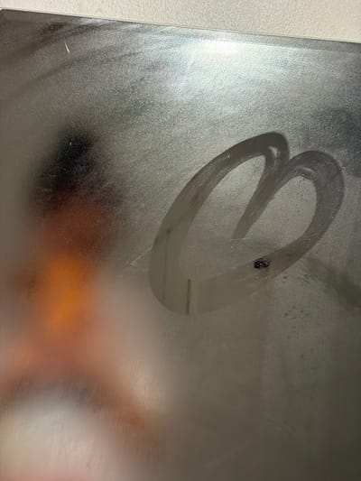 The image shows a drawing of a heart on a steamy glass or mirror surface. The heart is not perfectly formed and appears to have been drawn with a finger, leaving streaks in the condensation. A small black object, possibly a bug, is present inside the heart shape. The rest of the surface is covered in condensation, obscuring the background. It is impossible to pinpoint the exact location within Overstrand Local Municipality based on the given information. The image appears to be a close-up, focusing on the heart drawing and the surrounding condensation. There are no identifiable people, specific subjects, or actions occurring other than the drawing itself.