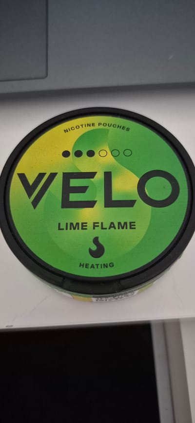 The media file shows a close-up, overhead shot of a round, black container with a green and yellow lid. The lid features bold, black text and symbols. Dominating the center of the lid is the word "VELO" in large, stylized letters. To the left of "VELO", smaller text reads "NICOTINE POUCHES" vertically. Below this, there are three black dots and two white circles arranged in a row, possibly indicating strength or flavor levels. To the right of "VELO", the text "LIME FLAME" is written vertically, followed by a stylized flame-like symbol, and then the word "HEATING" below it. The background is mostly out of focus, but appears to be a light-colored surface, possibly a table or desk. The lighting suggests an indoor setting, likely during the day given the brightness. There are no people or clear environmental cues to suggest a specific time of day or weather conditions beyond the indoor setting. The location context is Lillehammer, Norway, but this information is not visually discernible from the image itself. The overall impression is of a product shot, focusing on the branding and details of the container.