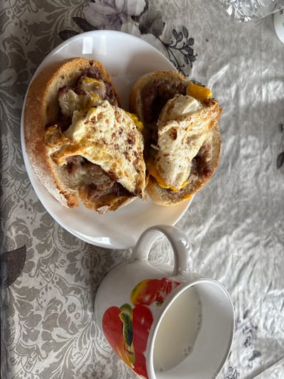 An overhead view captures a meal set on a patterned tablecloth in Kastel, Bulgaria. A white ceramic plate holds two open-faced sandwiches, each consisting of a halved bread roll. The rolls are filled with a dark, granular substance, consistent with meat or sausage, topped with melted yellow cheese and a fried egg exhibiting browned, crisped edges and visible dark specks of seasoning. To the lower right, a white ceramic mug with a handle, decorated with red and yellow fruit or vegetable images, contains a white liquid, consistent with milk, with small bubbles on its surface. The tablecloth features a repeating grey floral or damask pattern on a light background.