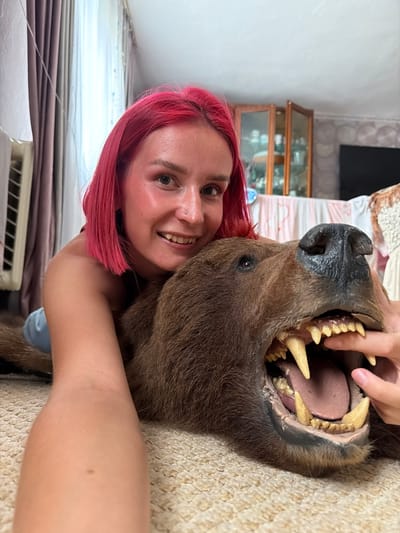 A young woman with bright pink hair is lying on the floor, resting her head on the open jaws of a taxidermied bear. Her right arm is extended towards the bear's mouth, with her finger touching its teeth. She is smiling at the camera, looking directly at the viewer. The bear's mouth is open wide, revealing sharp teeth and a pink tongue. The background shows a domestic interior with curtains, a cabinet with glass doors, and a television. The floor is covered with a light-colored carpet. This scene is situated in the city of Заболотово, Russia.
