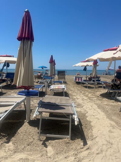 The image shows a beach in Bashkia Durrës, Albania. There are numerous beach umbrellas with red and white canopies and beach chairs arranged on the sandy beach. Some people are sitting in the chairs. The sea is visible in the background under a clear blue sky. The image is rotated.
