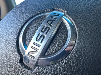 The image shows a close-up of the Nissan logo on a car's steering wheel. The logo is metallic and features the word "NISSAN" in raised lettering within a circular frame. The steering wheel itself is made of black textured leather. The lighting suggests it is daytime, with reflections visible on the metallic logo. There is no indication of the city of Shatava, Ukraine, in the image.