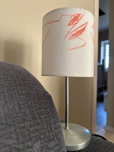 The image shows a lamp with a cylindrical white lampshade covered in red crayon markings. The lamp is attached to a grey upholstered object, possibly a piece of furniture. The supporting arm is a thin metal rod, connected to a circular metal base. The background is a blurred view of what appears to be an interior space with a light fixture visible in the distance. The location, Mežezeri, Latvia, cannot be determined from the image alone.