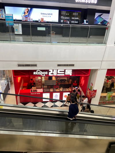 In the city of Kudus, Indonesia, a top-down view captures an escalator in a shopping mall. The escalator is moving upwards, with several individuals riding it. Directly ahead and slightly to the right, a brightly lit red kiosk with the signage "REDDOG" and Korean characters is visible. The kiosk appears to be a food stall, with people queuing to order or pick up their food. Behind the kiosk, a display refrigerator filled with food items and a counter area are discernible. The overall environment suggests a busy commercial space, likely within a mall.