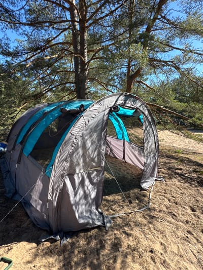 An unpitched tent is set up on a sandy, pine-forested area in Ozerki (Krasnodolinskaya), Russia. The tent is predominantly gray with turquoise accents on its upper sections. It is a dome-style tent with multiple entrances and mesh windows. The tent appears to be in a partially assembled state, with some fabric lying on the ground. The surrounding environment consists of tall pine trees with visible branches and needles, casting shadows on the sandy ground. The sky is a clear blue. The ground is covered in sand, pine needles, and dry grass. There are no people visible in the image.
