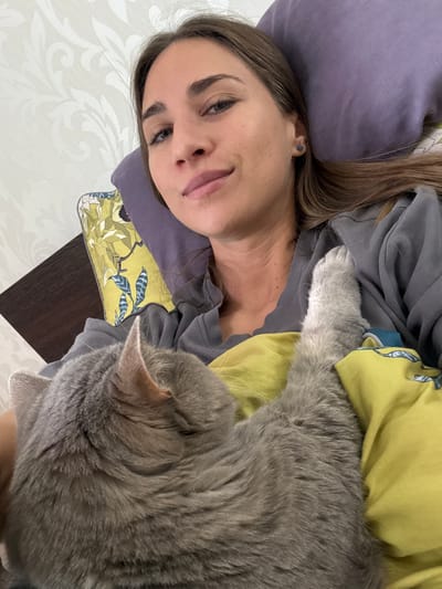 A woman lies on a bed with a gray cat. The woman has brown hair and is wearing a gray sweatshirt. She is resting her head on a purple pillow. The cat is lying on top of her, partially on a yellow pillow with a floral pattern. The bed is covered with a white sheet with a floral pattern. A dark brown surface is visible near the pillows. The location is Zanevka, Russia.
