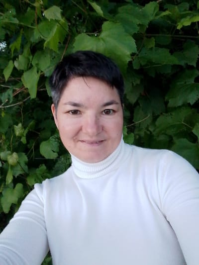 This is a selfie of a person with short, dark hair and brown eyes, wearing a white turtleneck sweater. They are smiling and looking directly at the camera. The background consists of lush green foliage, possibly from a vine or bush, with some smaller, lighter green clusters visible. The lighting suggests it could be daytime, and the natural background hints at an outdoor setting, possibly in a garden or park. The location context provided is Savino, Russia.
