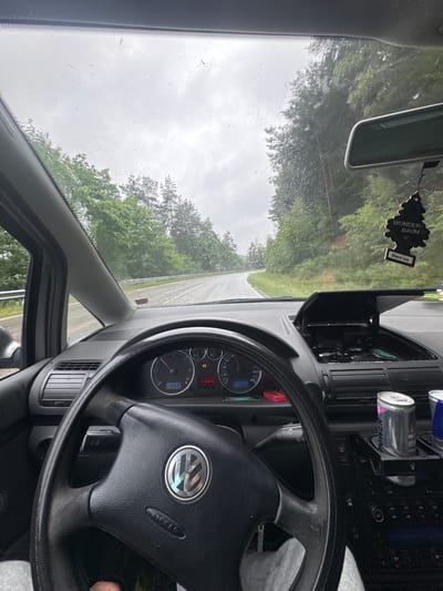 This media file captures the interior of a vehicle from a driver's perspective, looking out through the front windshield towards a road winding through a wooded area. The scene depicts a cloudy, overcast day, with visible raindrops on the windshield, suggesting recent or ongoing rain.
In the foreground, the black steering wheel of what appears to be a Volkswagen vehicle (indicated by the VW logo in the center) dominates the lower right portion of the frame. The word "AIRBAG" is visible on the steering wheel. To the left of the steering wheel, a dashboard instrument cluster shows a speedometer, a tachometer, and other gauges. A digital display reads "14.0°C" (likely the outside temperature) and "653" (possibly a trip odometer or time). Several warning lights are illuminated on the dashboard. To the right of the dashboard, air vents are visible, and further to the right, two drink cans are placed in a holder. One can clearly shows the text "POWER SUGARFREE" upside down.
Hanging from the rearview mirror is a "WUNDER-BAUM" (Little Trees) air freshener, labeled "Black Ice." The rearview mirror itself is visible in the upper center. The windshield is speckled with raindrops, and beyond it, a two-lane road is visible, flanked by dense green trees under a bright, cloudy sky. The road appears wet.
The overall impression is of a car parked or slowly moving on a rural or semi-rural road in Steinalben, Germany, during a cool, damp daytime.