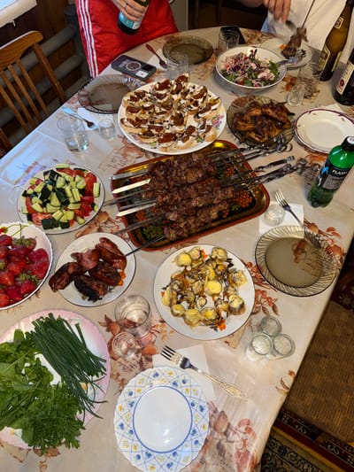 This image depicts a table laden with food and drinks, suggesting a gathering or meal. The setting appears to be indoors, possibly a home, with a patterned tablecloth covering the table. Several dishes are visible, including skewers of grilled meat, a salad, an assortment of appetizers on bread, fried eggs with some other food item, strawberries, sausages and grilled eggplant, and a plate of green vegetables and herbs. There are also several glasses, some containing clear liquid, suggesting alcoholic beverages like vodka. Bottles of drinks are also present, including a green bottle with a label. The presence of multiple plates and cutlery indicates that people are, or were, dining here. The overall impression is one of a shared meal with a variety of food and drink options.