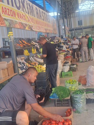 The image depicts an outdoor market stall, likely in Ko`kdala, Uzbekistan, filled with a variety of fresh produce. A man wearing a grey t-shirt and shorts is crouching in the foreground, reaching into a cardboard box filled with ripe red tomatoes. To his right, a black plastic crate contains more of these tomatoes. Behind him, a large pile of dill or fennel is displayed on a black crate, next to a clear plastic bag filled with green bell peppers and another bag containing cucumbers. In the background, other market-goers are visible browsing the stalls. Some men are seen standing near displays of fruits and vegetables, including bananas, apples, and onions. Numerous crates and bags of produce are stacked throughout the scene, indicating a busy marketplace. A large banner with text in a foreign language is displayed above the stalls. The overall environment suggests a typical agricultural market setting.