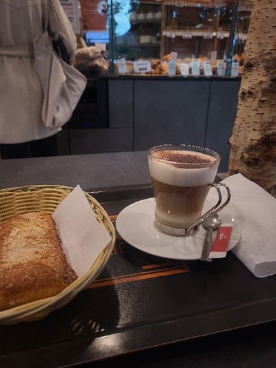 A close-up, first-person perspective shot depicts a table setup inside a bakery or cafe in Publier, France. The foreground is dominated by a dark rectangular serving tray. On the left side of the tray, a light-colored wicker basket contains a rectangular, golden-brown pastry with a crumble topping, accompanied by a folded white paper napkin. To the right of the basket, a clear glass mug with a metal handle holds a layered latte or cappuccino, featuring distinct layers of dark liquid, lighter liquid, and white foam, dusted with dark powder. This mug rests on a white ceramic saucer, which also holds a small silver-colored spoon and a red sugar sachet. A second folded white paper napkin is positioned to the right of the coffee setup on the tray. On the far right of the frame, a decorative section of a light-colored, textured tree trunk is vertically positioned. In the background, a person, visible from the chest down, wears a light-colored coat and carries a light-colored shoulder bag while standing near a long glass display counter filled with various baked goods, including loaves of bread and pastries, each with small white identification labels. The counter beneath the display is dark grey.