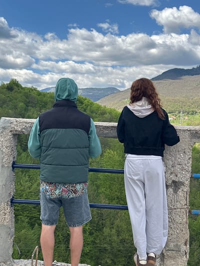 Two individuals are standing on a concrete structure overlooking a valley. The person on the left is wearing a black jacket over a white hoodie and grey pants, along with brown shoes. The person on the right is wearing a green hoodie and a vest with black and green sections, denim shorts, and tan skin. A blue railing is in front of them. The background features mountains, trees, and a cloudy sky. The location is Đurđevića Tara, Montenegro.