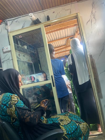 The image shows a reflective display case. A man in a blue shirt is lying down, possibly on a surface behind the case. Two people are reflected in the case, one holding a mobile phone. The setting seems to be indoors, with visible walls and a ceiling structure made of corrugated metal sheets. Given the reference to Okengwe - Obehira, Nigeria, the environment appears to be a modest commercial or residential space. There are electrical installations visible, suggesting a functional indoor area.