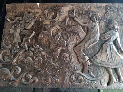 This is a detailed, close-up, eye-level photograph of a large, carved wooden relief panel depicting a lively scene. The carving is in a rustic, folk art style, with figures and decorative motifs raised from a textured background.
On the left side of the panel, a figure wearing a hat and a scarf is seated and playing a stringed instrument that resembles a guitar or lute. The figure is dressed in clothing with prominent folds and appears to be in motion, with one leg bent and the other extended.
To the right of the musician, a larger male figure, also wearing a hat, is shown in a dynamic pose, with one arm raised and his body leaning back. He appears to be dancing or celebrating. Next to him, a female figure in a long dress is also depicted in a dancing posture, with her arms at her sides.
The figures are surrounded by elaborate, swirling floral and vine-like patterns that fill the negative space. These decorative elements are carved with depth and detail, creating a sense of movement and organic growth.
The wood has a dark, weathered finish, with visible grain and texture. The carving is deep, creating strong shadows and highlights that emphasize the three-dimensional quality of the artwork.
The context of Hovden i Setesdal, Norway, suggests this carving might be a representation of traditional Norwegian folk culture, music, and dance.