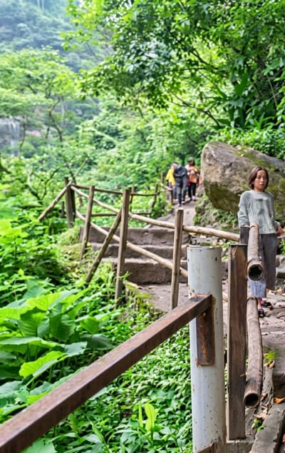 A young woman stands on a stone path next to a rustic wooden fence and a metal railing, looking forward with a neutral expression. She is wearing a light gray, long-sleeved sweater and gray pants. In the background, a group of people walk up a stone staircase, surrounded by lush green foliage and trees. The scene appears to be a nature trail in a mountainous or hilly area, likely in Dolat Rayat, Indonesia, given the tropical vegetation.