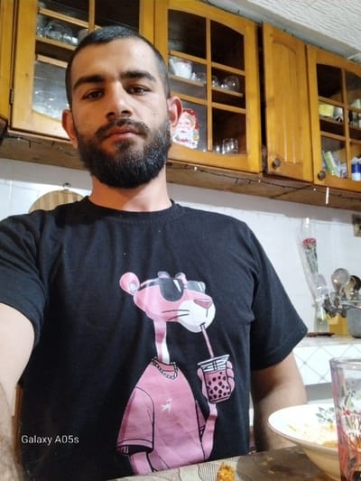 An adult male is captured in a headshot/upper torso photograph within a residential kitchen or dining area in Oreshene, Bulgaria. He has dark hair, a full dark beard, and is looking directly at the camera. He wears a black t-shirt featuring a cartoon character resembling the Pink Panther, depicted wearing sunglasses, a chain, and a pink shirt, while drinking from a cup with a straw. The background includes light brown wooden wall cabinets with glass inserts, showcasing various clear glassware and ceramic items. A string of small white lights is affixed beneath the cabinets, and a decorative Santa Claus face is visible within one cabinet. To the right, kitchen utensils are partially visible on a counter. In the foreground, a white bowl containing food residue and a clear glass with a red pattern holding a beverage are present on a patterned tablecloth. A "Galaxy A05s" watermark is present in the lower-left corner of the image.