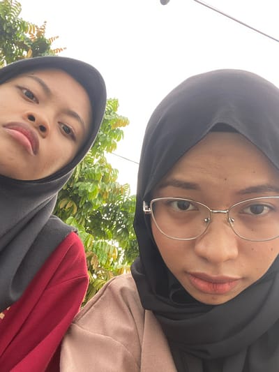 This is a close-up, selfie-style photograph featuring two individuals, likely female, wearing hijabs. The upper subject is in the foreground and is wearing glasses with thin, round metal frames. Her head is tilted, and she is looking directly at the camera. Her lips are slightly parted, and her skin appears to have some texture. The second subject is positioned below and to the left of the first, also wearing a hijab. Her face is angled away from the camera, with only her eyes and mouth clearly visible. She has a neutral expression.
Both individuals are wearing different colored tops; the upper subject has a light pink or beige garment visible on her shoulder, while the lower subject is wearing a dark gray hijab and a dark red top. The background is predominantly green foliage, suggesting an outdoor setting with trees or bushes. The sky is overcast and bright, indicating it is daytime. There is a thin, metallic pole visible in the upper left portion of the image, extending upwards. No text is visible in the photograph. The image appears to be taken in a casual, everyday setting, possibly a park or a backyard, given the presence of greenery and the casual attire. The overall mood is informal and candid. The location context is Air Molek, Indonesia.