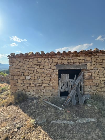 The image depicts a small, dilapidated stone structure with a terracotta tile roof situated on a hillside. The structure has a large, open doorway filled with broken wooden debris, suggesting disrepair. The surrounding environment is dry and arid, with sparse vegetation and loose stones. In the background, a hazy landscape of hills and distant mountains is visible under a clear blue sky with scattered clouds. The sun is shining brightly, casting shadows across the ground. The style of the stonework and roofing is consistent with traditional rural architecture found in the region of Alpuente, Spain.