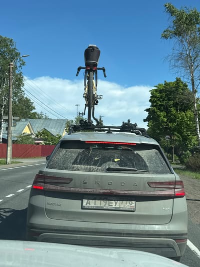 The image depicts the rear view of a dirty grey Skoda Kodiaq SUV traveling on an asphalt road under a clear blue sky with scattered white clouds, in Beloostrov, Saint Petersburg, Russia. A yellow-framed bicycle with drop handlebars and a black saddlebag is mounted vertically on a roof rack on top of the vehicle, facing backward. The car's tailgate and rear window are heavily splashed with dirt and mud. The rear taillights are illuminated red. The Russian license plate, "A 119 EY 178," includes the region code "178 RUS" (Saint Petersburg) and text indicating "Авто Премиум" and "СПб. пр. Энгельса 15" below it. The car also features a "KODIAQ" badge and a "4x4" badge on its tailgate.

To the left of the vehicle, a wooden utility pole with a street light and multiple overhead power lines stands beside the road. Beyond the pole, a red wooden fence partially obscures two houses with grey roofs, one of which has yellow walls. Deciduous trees with light-colored bark are visible on both sides of the road. To the right of the vehicle, a circular white sign with a red border displays "40" in black, indicating a 40 km/h speed limit. The road features white dashed lane markings. Bright sunlight suggests a daytime scene. No people are directly visible.