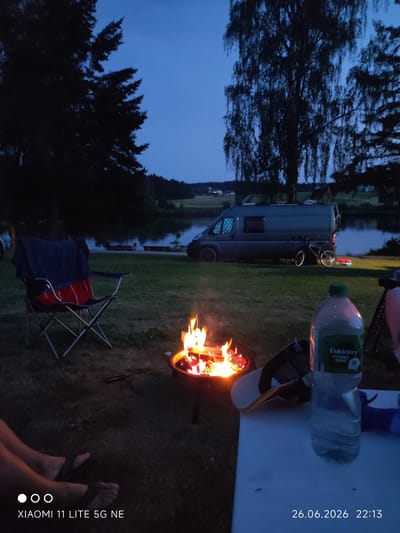 The image captures a serene evening scene at a campsite, likely near Abtsgmünd, Germany. The time is dusk, as indicated by the deep blue of the sky and the presence of a lit campfire. The setting is a grassy area next to a tranquil body of water, possibly a lake or river, with trees framing the background.

In the foreground, a portable campfire crackles with bright flames, casting a warm glow. To the left of the fire, a folded camping chair is set up, draped with a dark blue cloth and a red item. Further left, a person's legs and feet, wearing flip-flops, are visible, suggesting relaxation and informality.

Dominating the mid-ground is a grey camper van, parked on the grass. A bicycle leans against the side of the van, hinting at outdoor activities. In the right foreground, a partially filled plastic bottle of water sits on a white surface, possibly a picnic table. The label on the bottle is partially visible, with "Volvic" being legible.

The overall atmosphere is peaceful and relaxing, embodying the essence of a camping trip. The soft lighting from the campfire and the twilight sky contribute to a cozy and calm mood. Visible text on the image includes the camera information "XIAOMI 11 LITE 5G NE" and the date and time "26.06.2026 22:13". The presence of a body of water and the type of vegetation suggest a natural, possibly rural, environment.