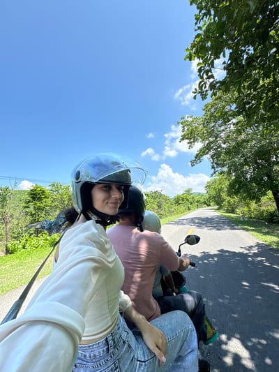 This is a first-person perspective photograph taken from the back of a motorcycle, traveling on a paved road. The road is flanked by lush green vegetation and trees, with a clear blue sky overhead dotted with white clouds. In the foreground, a young woman wearing a white top, blue jeans, and a clear helmet is visible. She is looking at the camera and smiling. Behind her, two other people, one wearing a pink shirt and the other a gray helmet, are also on the motorcycle. The scene suggests a casual outdoor activity, possibly a scenic drive. The location is the city of Hàm Thuận Bắc, Vietnam.