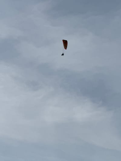 A single paramotorist is captured in mid-flight against a cloudy, overcast sky in Žemieji Rusokai, Lithuania. The paramotor, an orange and yellow wing, is positioned in the upper center of the frame, with a silhouetted figure piloting it. The clouds are dense and gray, covering the entire sky and obscuring any clear visibility of the horizon or landscape below. The overall atmosphere is somber and muted due to the lack of sunlight.