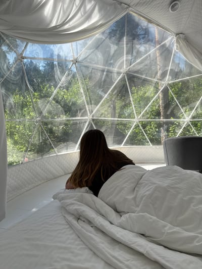 The image depicts the interior of a geodesic dome, likely a glamping accommodation in Nechkino, Russia. A person with long brown hair is lying on a bed covered with white bedding, facing away from the camera and looking out through the transparent dome. The dome's structure, composed of white geometric frames, is visible. Outside the dome, a lush green forest and blue sky are visible. The interior features white padded walls and a white floor. A dark grey armchair is partially visible in the background. The lighting suggests it is daytime.