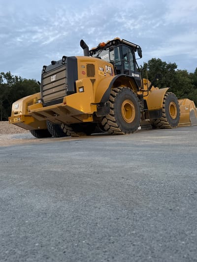 A large yellow Caterpillar 962K wheel loader is parked on a paved surface, likely in a construction or industrial area. The loader is viewed from a low-angle, ground-level perspective, emphasizing its size and robust construction. The front of the loader, including the iconic CAT logo on the grille, is prominently featured. The machine's large tires, heavy-duty bucket, and exhaust stack are visible. The background consists of a line of trees and a cloudy sky. There are no people or obvious signs of recent activity in the image. The location is specified as being in Grenay, France, though no specific landmarks are identifiable within the frame that would confirm this location.
