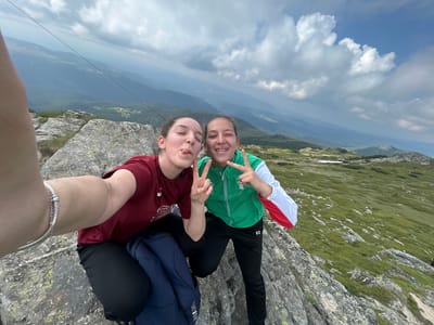 Two young women are taking a selfie on a rocky mountain ledge. The woman on the left is wearing a maroon t-shirt and black pants. She is sticking her tongue out at the camera. The woman on the right is wearing a green and white jacket and black pants. She is making a peace sign with her right hand. Both women are smiling. In the background, there is a vast mountain landscape with rolling hills and cloudy skies. The location is likely in the vicinity of Cherna Mesta, Bulgaria, given the mountainous terrain and vegetation.
