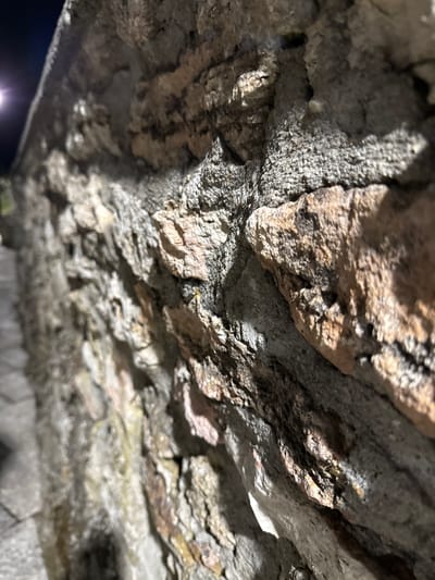The image is a close-up, low-angle shot of a textured stone wall, likely part of a building or an outdoor structure in Tyulenovo, Bulgaria. The wall is constructed from rough, irregularly shaped stones of varying sizes and colors, primarily grays, browns, and some reddish hues. The stones are bound together with a coarse, cement-like mortar, also gray in color. The surface of the stones and mortar is rough and uneven, with visible pores and crevices.
In the upper left corner of the image, a faint, blurred light source is visible, possibly a street lamp or moonlight, casting shadows and highlighting the texture of the wall. The background is dark and out of focus, suggesting it's nighttime. The perspective is angled, with the wall extending from the foreground towards the background and receding to the left. The ground, made of paving stones, is visible along the lower left edge of the image, also out of focus.
There are no people or animals in the frame. The focus is entirely on the architectural details of the stone wall. The overall impression is one of ruggedness and age.