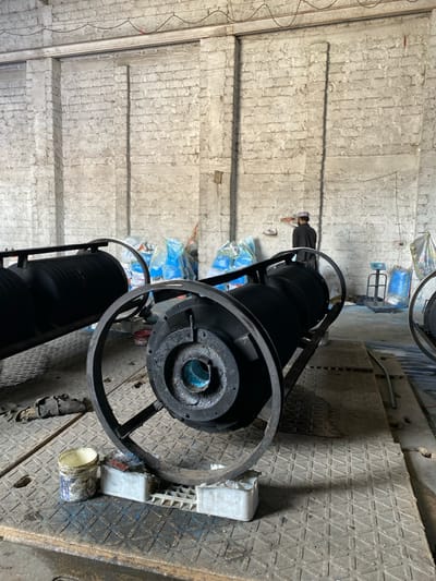 The image depicts an indoor industrial or workshop setting located in Daman, Afghanistan. In the foreground, two large, dark, horizontally oriented cylindrical objects are present, each encased within a black metal frame with prominent circular end supports. The closest object, resting on two white plastic crates, displays a central circular opening with a light blue interior. A dirty white plastic bucket with blue residue sits on the floor nearby. The floor is covered with dark, textured metal plates showing signs of wear and blue staining. In the mid-background, a male individual is observed standing with his back to the camera, wearing dark clothing and a light-colored cap. He appears to be observing or interacting with an object on the rough, light-colored brick wall. Behind him, against the wall, are stacks of blue and white plastic bags, and to the right, an industrial platform scale is visible with a blue bag on or near it. The walls are constructed of irregular light-colored bricks with vertical concrete or plaster elements.
