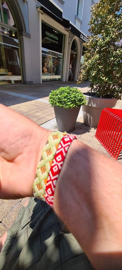 This media file shows a close-up view of a person's arm wearing two woven bracelets, one red and white with a diamond pattern, and the other green, orange, and white with a crosshatch pattern. The bracelets are made of thick thread, suggesting they are handmade friendship bracelets. The person is wearing green plaid shorts.
In the background, there is a storefront with a black awning that reads "CALZEDONIA" in white letters. Below this, the storefront window displays mannequins wearing undergarments, and the words "CALZEDONIA Intimissimi" are visible on the window. Further down, another storefront has the word "GUESS" visible in large letters.
To the left of the storefront, there is a large grey planter with a lush green bush. Next to it, a bright red, slatted chair is partially visible. The ground is paved with light-colored rectangular tiles. Sunlight casts distinct shadows, indicating it is daytime. The scene appears to be an outdoor shopping area, likely a pedestrian street in a town or city. The overall impression is sunny and casual.
There is no clear indication of the emotion of the person, beyond the casual attire and the presence of friendship bracelets, which might suggest a relaxed or friendly mood. The context of the location being Aiello del Friuli / Dael, Italy, suggests a European setting.