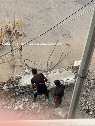 A top-down view shows two men engaged in construction work near a roadside in an unidentified area. The men are dressed in dark clothing, with one visible wearing black pants with white stripes and sandals. They are working with rebar and construction debris, which are scattered around them. The location appears to be urban, evidenced by the concrete road surface and a utility pole overhead. The photograph captures aspects consistent with construction-related activities in a populated setting. It is impossible to determine the city of al-Qarara, Palestinian Territories location from the image provided.