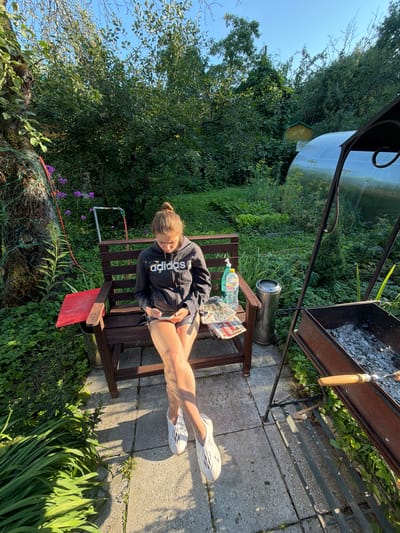 A young woman with brown hair in a bun, wearing a black Adidas hoodie and grey shorts, sits on a wooden bench in a backyard. She is looking down at her phone, which she holds in her hands. To her left, a red wooden tray is attached to the bench. To her right, a plastic bottle of water and a small pile of magazines rest on the bench. A silver trash can is located behind the bench. In the background, there are lush green trees, a greenhouse, and a grill. The scene is set in the city of Чайка Снт (купавна Старая), Russia, bathed in the warm light of the sun.