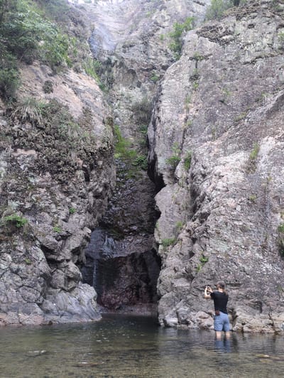 An outdoor scene in Godang-ri, South Korea, displays a deep, natural gorge featuring steep, rugged rock faces that rise vertically on both sides of a shallow, clear body of water. The rock surfaces are predominantly grey and brown, exhibiting varied textures and geological strata, with sparse patches of green vegetation, including moss, grasses, and small shrubs, clinging to cracks and ledges. On the left side of the frame, at water level, a dark, cavernous opening is visible, with a small waterfall flowing into it. The water at the bottom reveals a rocky bed through its transparency and shows minor surface ripples and some foam. In the lower right quadrant, a male individual stands in the water, which reaches his mid-thigh. He is facing the left side of the gorge, wearing a black t-shirt and light blue shorts. He holds a dark electronic device, resembling a smartphone or small camera, with both hands, positioned as if capturing an image or video of the cave and waterfall area. A small, brown leather pouch is affixed to his right hip.