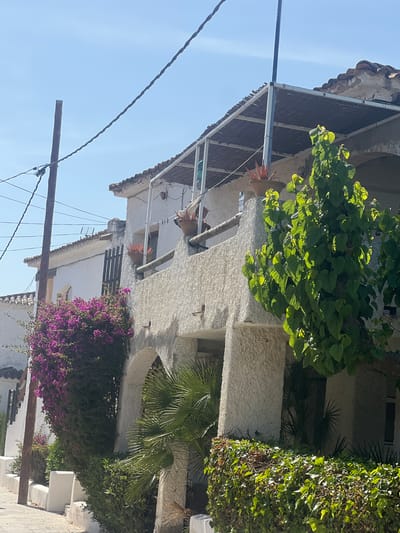 The image depicts a two-story building located in La Marina De Creixell, Spain. The building has a white, textured exterior with a tiled roof. Lush green vegetation, including bougainvillea with vibrant purple flowers, is growing around the building. Electrical wires are visible in the foreground, running diagonally across the blue sky. The architecture and vegetation suggest a Mediterranean environment.