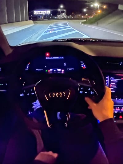 The image depicts the interior of a car at night, with the driver's perspective focused on the steering wheel and dashboard. The steering wheel, with the Audi logo illuminated in orange, is partially obscured by a hand. The dashboard displays a variety of illuminated readouts, including speed, gear selection, and a navigation interface. Outside the car, the road is visible, marked with blue and white lines, indicating a highway or major road. In the distance, illuminated signs for "RHENUS LOGISTICS" are visible, suggesting an industrial or logistics hub. Streetlights and building lights dot the background, illuminating the surrounding environment. The scene appears to be in Bezden, Bulgaria, based on the context provided.