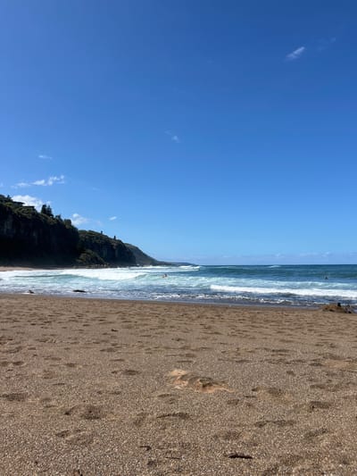 The image displays a daytime coastal landscape at Coalcliff, Australia, under a clear blue sky. Approximately the top half of the frame is dominated by the bright blue sky, exhibiting only a few small, scattered white cloud formations. The middle ground features the ocean, characterized by blue-green water with multiple parallel lines of breaking white waves extending across the scene.
On the left side of the ocean, a solitary individual is visible on a surfboard, positioned prone or paddling within a breaking wave. A second, less distinct figure is present further out in the water towards the right. The foreground consists of a broad expanse of damp, light brown granular sand, heavily marked with numerous human footprints. Irregular, shallow mounded formations and a small, lighter-colored sandy elevation are also present in the immediate foreground.
Along the left side of the image, a steep, dark cliff face, topped with dense green vegetation, rises from the water's edge and extends into the distance. Several white architectural structures, likely residential buildings, are visible on the crest of this vegetated cliff. Dark rocks are present at the interface where the waves meet the base of the cliff on the left, and a small cluster of similar dark rocks is visible on the far right edge of the sandy beach. The scene is illuminated by direct sunlight.