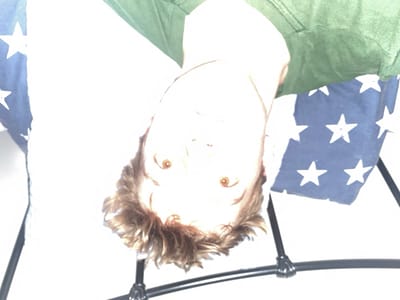 A person, likely a young male with blond hair, is hanging upside down with the top of their body, including their face, obscured by a white object, possibly a sheet or blanket. The person appears to be wearing a green shirt. The person's feet are out of frame, and their body is positioned above a black metal frame, which is obscured by an American flag in the background. The flag is positioned in the upper right portion of the image, with white stars on a blue background, and is draped over the metal frame. The image appears to be taken inside, with a white wall in the background. The image was taken at an unknown location, the coordinates provided for the city of Telford in the United Kingdom do not provide any relevant context for the image.