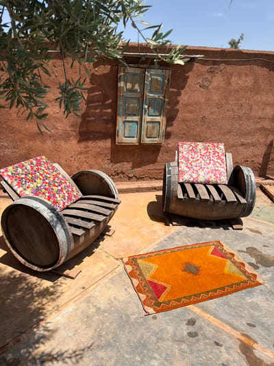 This image captures a vibrant and rustic outdoor seating area, likely in Tahannaout, Morocco, under bright sunshine. The scene is dominated by a textured, reddish-brown earthen wall, possibly adobe or mud brick, which shows signs of weathering and cracking.
In the foreground and midground, two large wooden barrels have been creatively repurposed as seats. Each barrel is cut in half horizontally, with wooden slats forming a backrest, and is adorned with a brightly patterned cushion. One cushion features a colorful floral design with red, pink, yellow, and purple hues, while the other appears similar.
On the greyish concrete or stone ground to the right, a rectangular rug with a warm color palette is spread out. It displays geometric patterns in shades of orange, yellow, red, and dark brown, characteristic of traditional Moroccan textiles. Strong shadows cast by the barrels and overhead foliage indicate bright sunlight, suggesting it is midday or early afternoon.
To the left, olive tree branches with silvery-green leaves partially obscure the view, adding a natural element to the composition. Higher up on the earthen wall, a small, faded wooden window or shutter structure, painted in a distressed blue, adds a touch of aged charm. A thin cable or wire runs along the top edge of the wall, where a small green plant also grows. The sky, clear and blue, is visible in the upper left corner. No people are present, and there is no visible text. The overall atmosphere is inviting, peaceful, and distinctly North African.