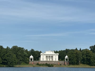 This is a landscape photograph featuring a large, white, neoclassical manor house with a portico of columns, situated on the shore of a lake. The manor is flanked by two white, domed gazebos, one on each side, atop a stone foundation. A dense forest of green trees forms the background behind the manor and gazebos. The foreground consists of a body of dark blue water, likely a lake, with reeds growing along the shoreline. The sky above is a clear, pale blue with some wispy white clouds. Two small figures in a boat are visible on the water in the lower left portion of the image. The manor house appears to be the Užutrakis Manor, located in Trakai, Lithuania, overlooking Lake Skaisčio.