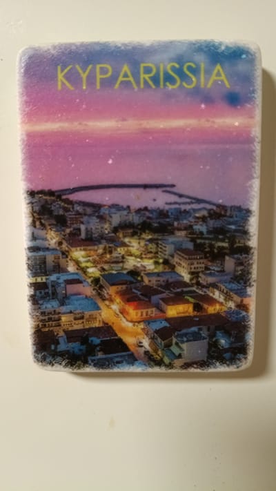 The image is a close-up of a souvenir magnet. The magnet features a photograph of a city at dusk or night, with a body of water in the background and a sunset or sunrise visible in the sky. The text "KYPARISSIΑ" is printed in yellow capital letters across the top of the magnet, indicating the location. The city depicted appears to be Kyparissia, a town in Messenia, Peloponnese, Greece. The photograph shows buildings with illuminated windows, suggesting an urban environment. The edges of the magnet have a slightly distressed or decorative border.