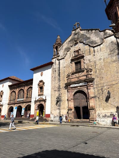 A vertical outdoor shot captures a building's facade under a clear blue sky. The architecture features aged stone, a prominent arched wooden door, and decorative window frames. People are visible on the steps and sidewalk, adding a sense of scale. Cobblestone paving and shadows indicate the time of day, possibly late morning.