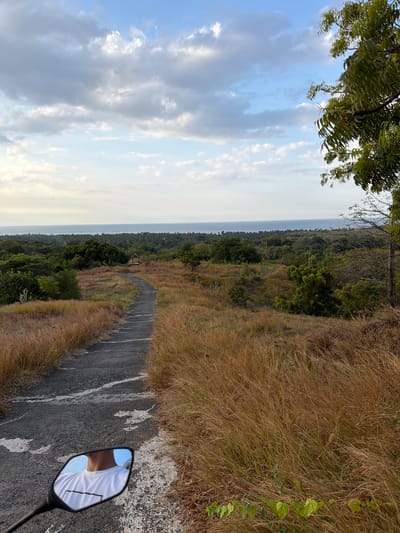 The image captures a scenic view of a paved road winding through dry, golden grass and sparse trees, leading towards the ocean horizon. A small, open-sided shelter is visible in the middle distance, nestled among the greenery. The sky is a mix of blue with scattered, light gray clouds. In the foreground, a motorcycle's side mirror reflects a person wearing a white t-shirt with a dark rectangular graphic. The scene suggests an outdoor location, possibly a viewpoint or a rural area on the outskirts of Kubutambahan, Indonesia, given the vegetation and coastal proximity. The lighting indicates it is either early morning or late afternoon.